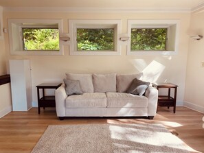 A living room with a beige sofa, two side tables, a wooden floor, and three windows with a view of greenery.