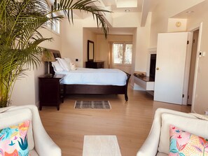 A bedroom with a bed, bedside tables, a nightstand, a large potted plant, and a view of the outside.