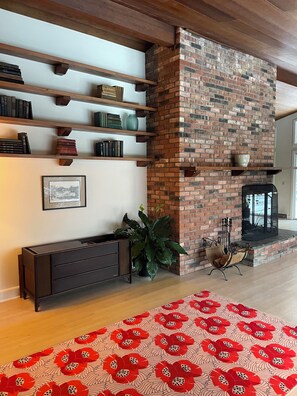 A living room with a brick fireplace, wooden shelves, a dark console table, a large potted plant, and a red patterned rug.