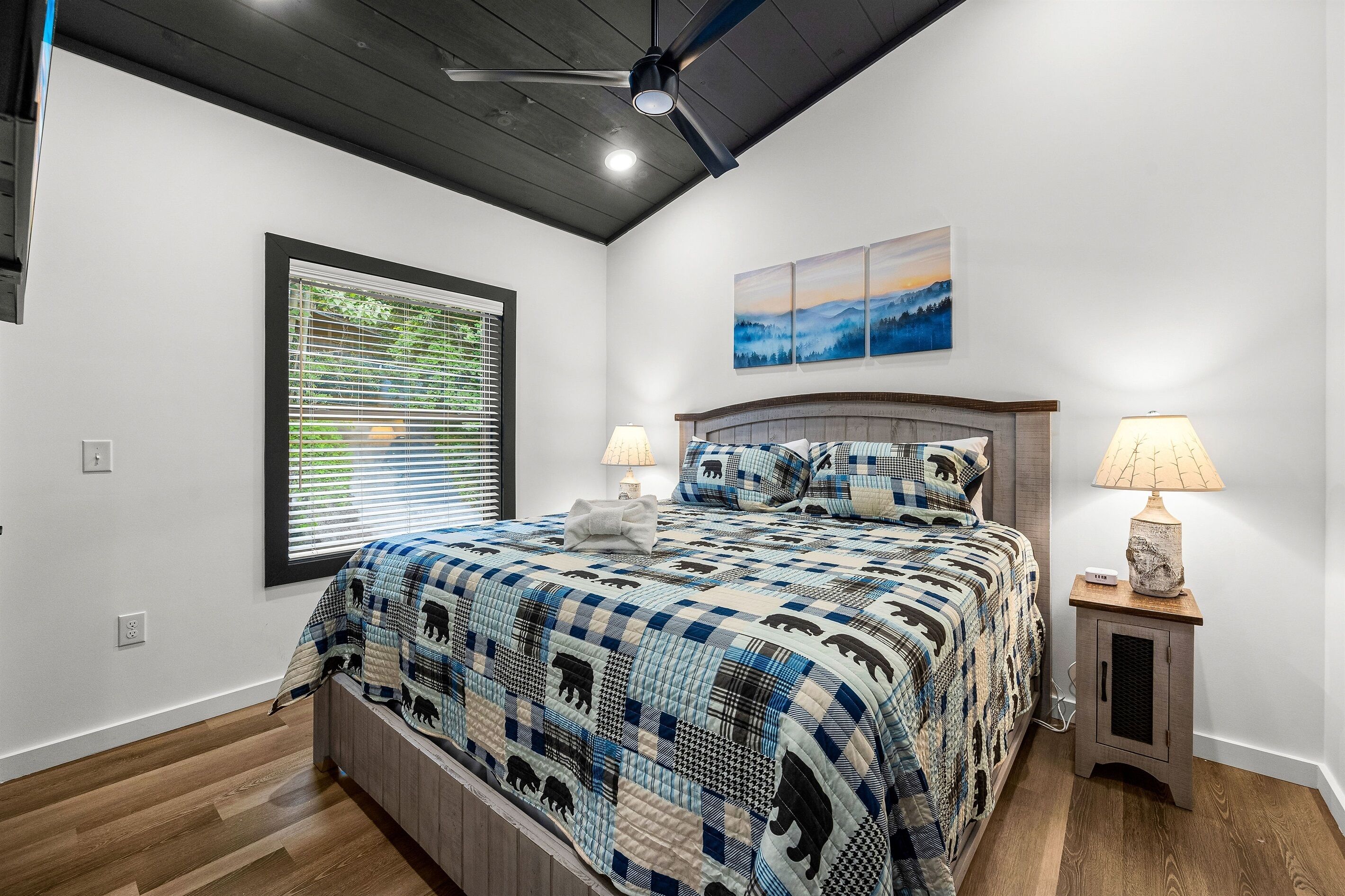 A bedroom with a bed, a window with blinds, a ceiling fan, a nightstand with a lamp, and a wall-mounted painting.