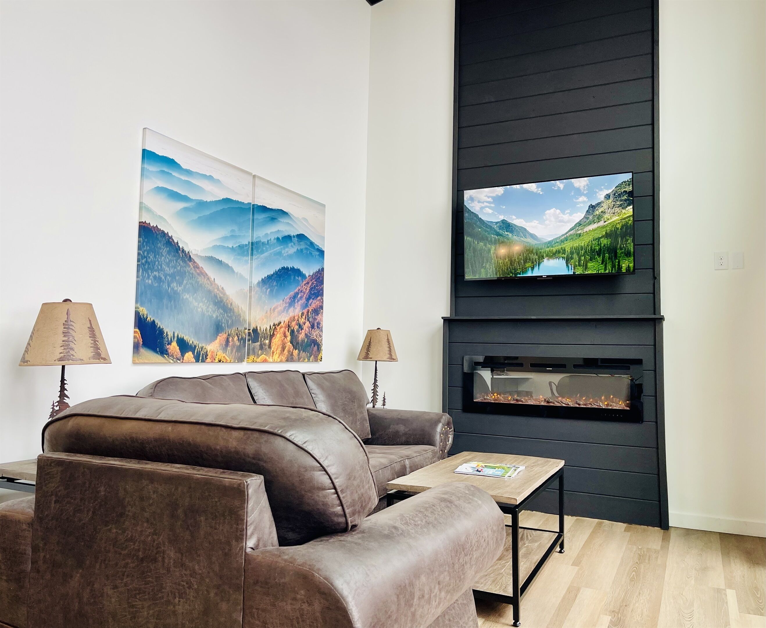Una sala de estar moderna con un sofá de cuero marrón, un televisor de pantalla plana sobre una chimenea y dos grandes cuadros de paisajes en la pared.
