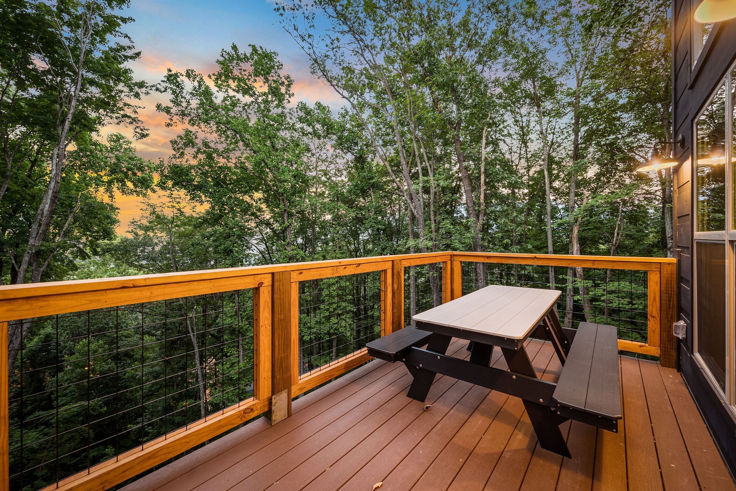 Une terrasse en bois avec une table de pique-nique et une rambarde donnant sur une zone boisée.