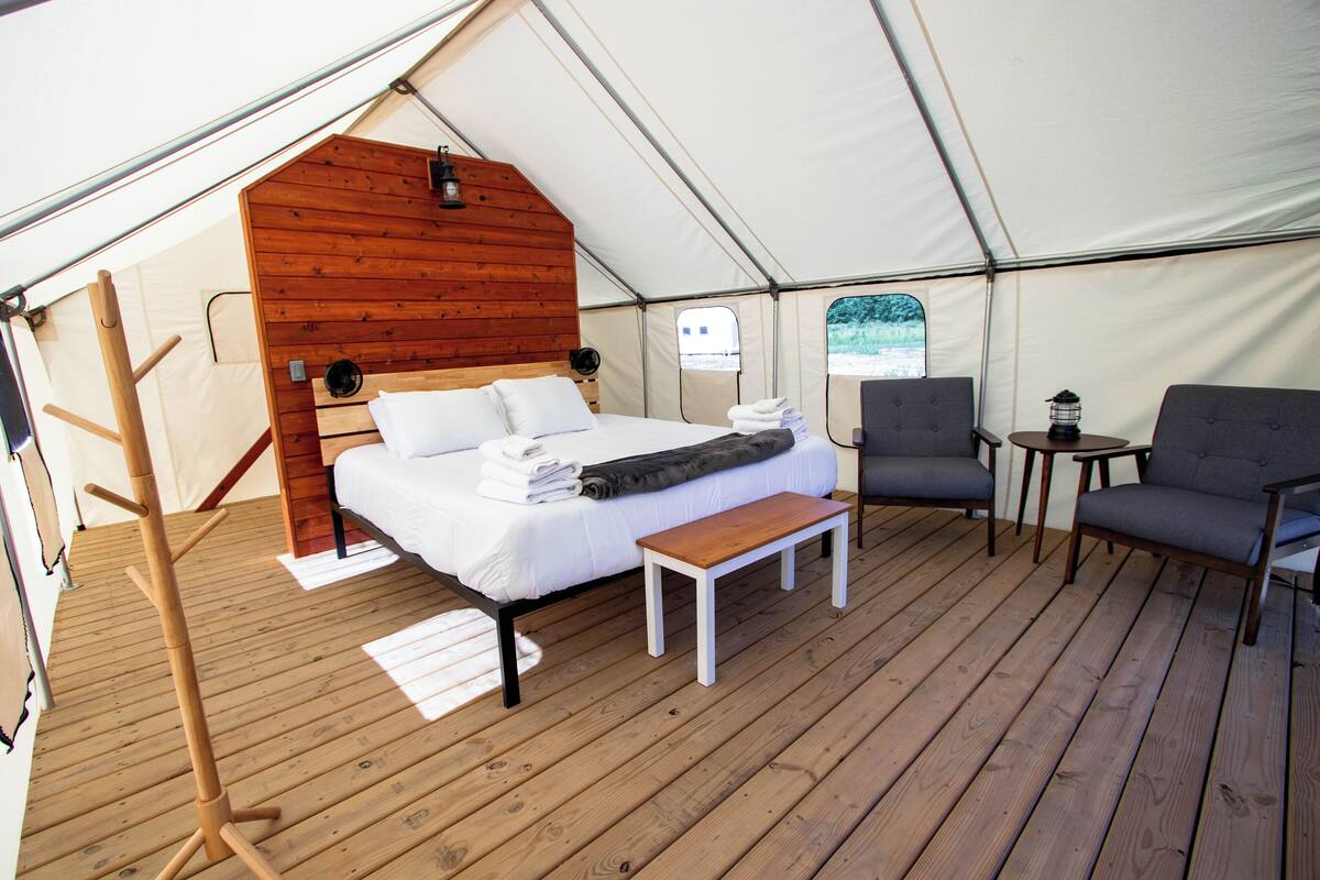 Eine Zelteinrichtung mit einem hölzernen Bett, einer Bank, einem Sessel und einem kleinen Tisch.