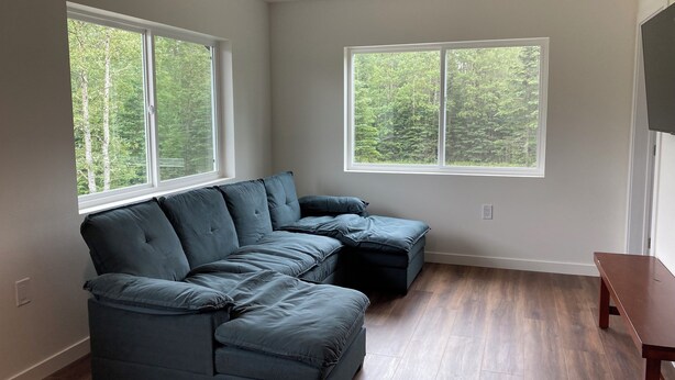 A modern living room with a sectional sofa, large windows with a forest view, and wooden flooring.