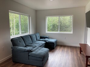 A modern living room with a sectional sofa, large windows with a forest view, and wooden flooring.