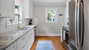 Una cocina con armarios blancos, un refrigerador de acero inoxidable, un lavamanos y una ventana con vistas a la vegetación.