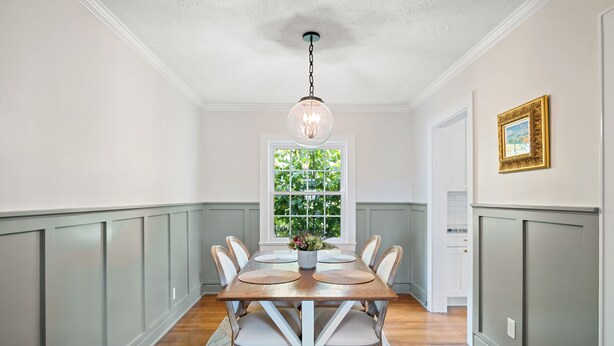 Comedor con mesa de madera, sillas blancas, una lámpara de araña y una ventana con follaje en el exterior.