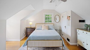 Un dormitorio con techo inclinado, una cama con cubrecama acolchado, una mesita de noche con lámpara, un aparador y un ventilador de techo.