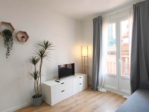 Une pièce avec un téléviseur posé sur un meuble blanc, deux plantes en pot, une lampe sur pied et une fenêtre avec des rideaux.