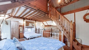 Une chambre à coucher au style rustique, avec un plafond de type «loft», un escalier en bois, un lit recouvert d’une couette à motifs floraux, une commode et un mur en pierre.
