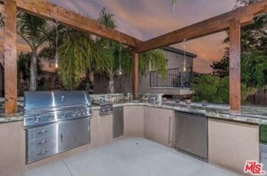 Une cuisine extérieure dotée d’un barbecue en acier inoxydable, de meubles de rangement intégrés et d’une structure de pergola en bois.