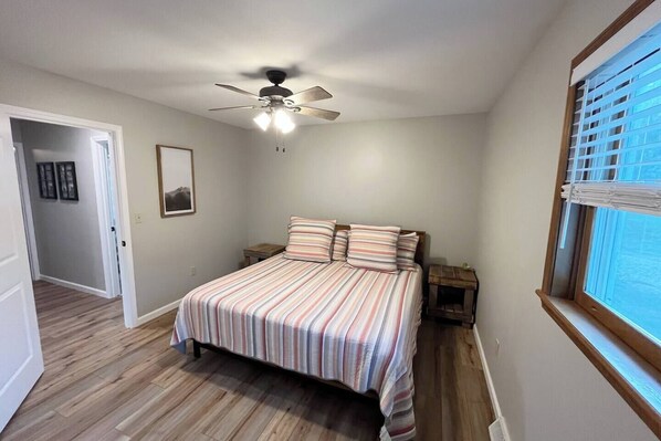 Un dormitorio con cama, ventilador de techo, ventana con persianas y puerta que da a otra habitación.