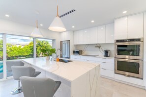 A modern kitchen with white cabinets, a large island with bar stools, stainless steel appliances, and a view of greenery outside. - Modern Oasis, Heated Pool, Short Drive to Beach (Fort Lauderdale)