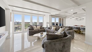 A spacious living room with large windows, a flat-screen TV, and a dining area with a table and chairs.