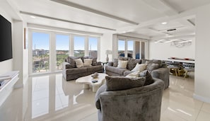 A spacious living room with large windows, a flat-screen TV, and a dining area with a table and chairs.