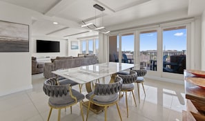 A spacious living area with a large dining table, grey upholstered chairs, a sofa set, and a flat-screen TV.