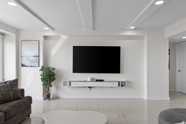 A modern living room with a flat-screen TV mounted on the wall, a white console table below, a grey sofa, a white coffee table, and a potted plant.