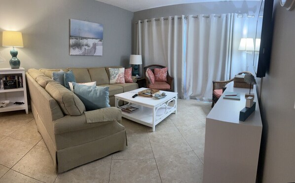 A living room with a beige sofa, a white coffee table, a bookshelf, a painting on the wall, and a window with curtains. - King bed, downstairs, 2/2, updated and spotless! (St. Simons Island)