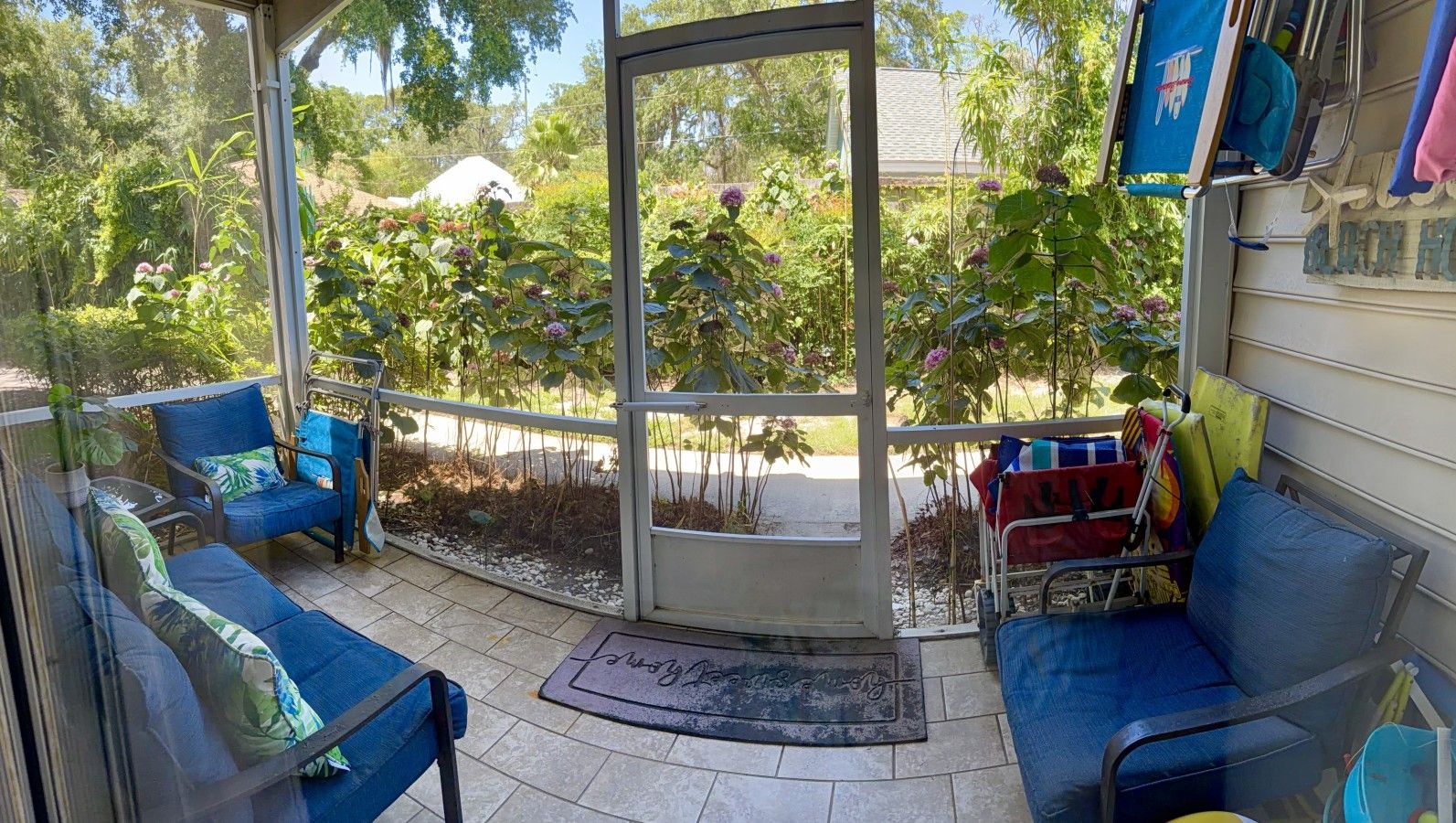 A covered outdoor seating area with two blue chairs and a bench, a glass door leading to a garden, and a small cart with cleaning supplies.