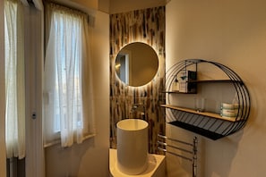 A bathroom with a round mirror, a cylindrical sink, and a shelf with toiletries. - Skyline-Bluemoon (Studio Apartment with a View) (Siena)