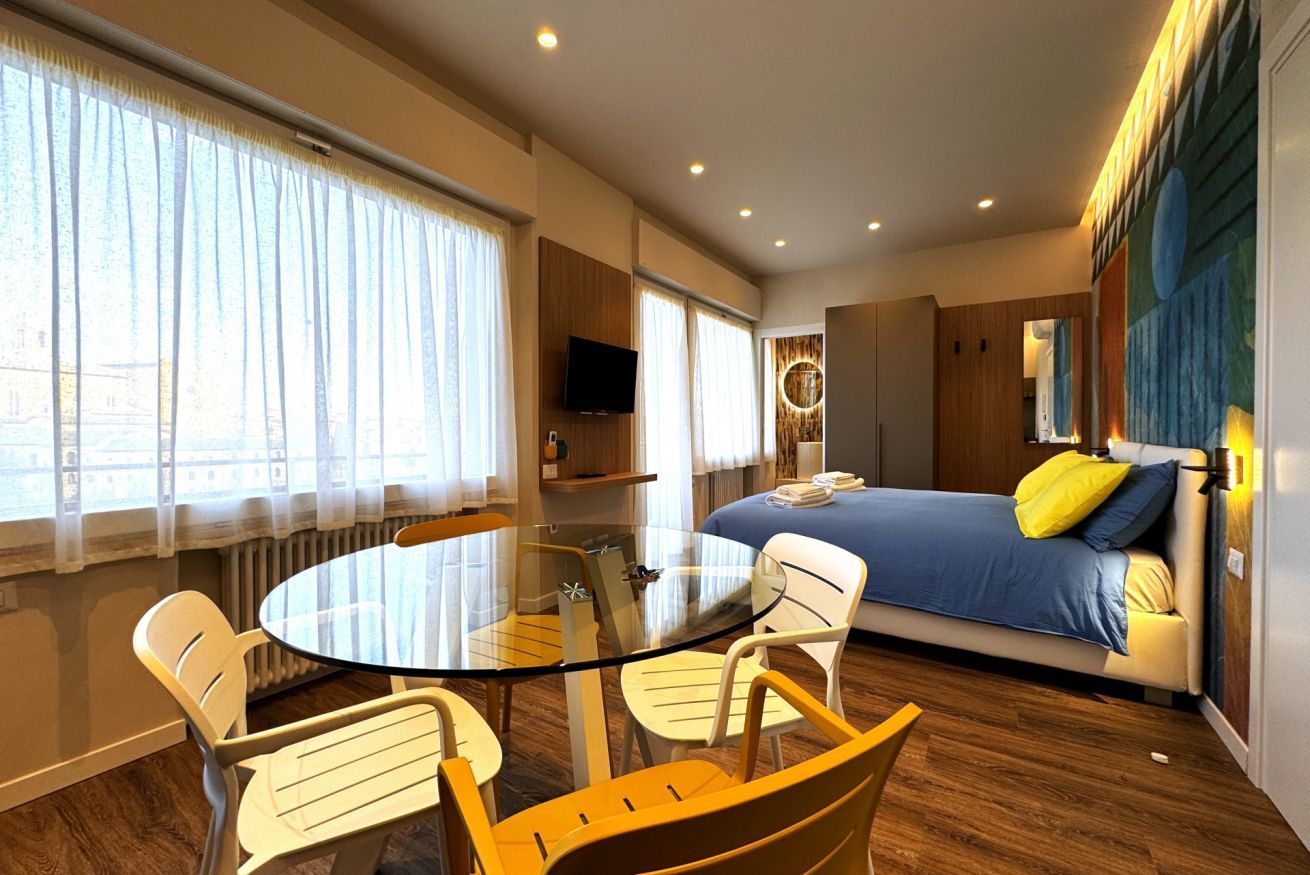 Een moderne hotelkamer met een ronde glazen tafel, witte stoelen, een bed met blauwe en gele kussens, een televisie en een groot raam met gordijnen.