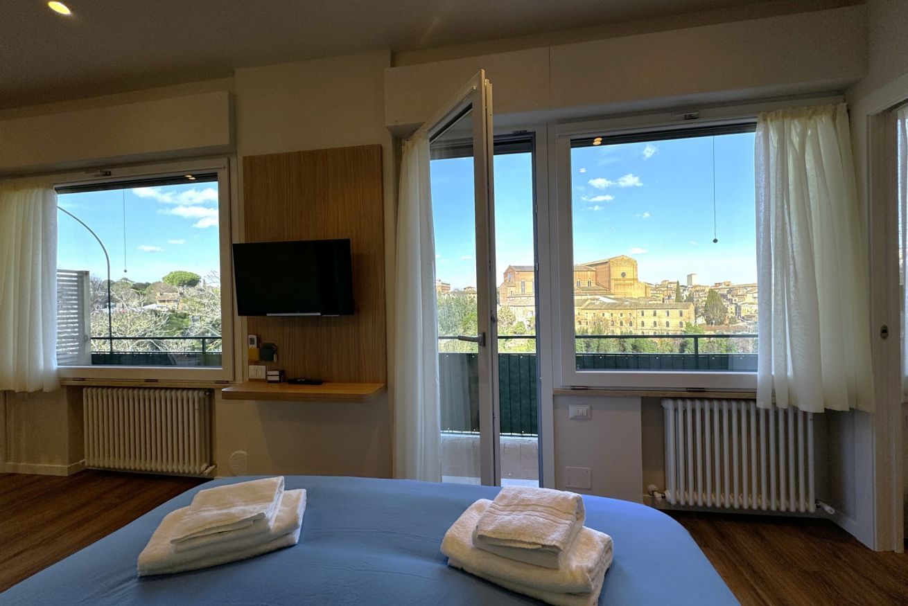 Habitación de hotel con cama, televisor montado en la pared y grandes ventanales con vistas a la ciudad.