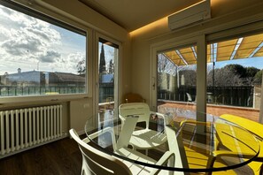A room with a glass table and chairs, a radiator, and large windows with a view of buildings and trees. - Skyline-Moncheri (Studio Apartment with a View) (Siena)