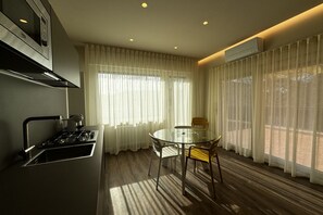 A modern kitchen with a gas stove, a glass dining table, and chairs. There is a window with sheer curtains and an air conditioning unit on the wall. - Skyline-Moncheri (Studio Apartment with a View) (Siena)
