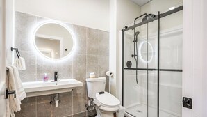 A modern bathroom with a glass shower enclosure, a round mirror with lighting, a toilet, and a sink with a towel rack. - Melwood Suites Unit 105 Studio (Pittsburgh)