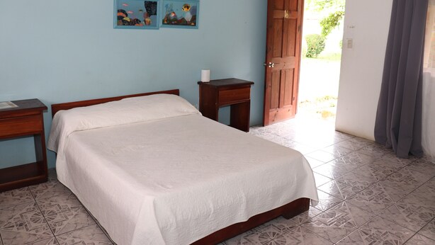 A bedroom with a bed, a nightstand, a small table, and a door leading outside. - Hotel Samoa del Sur (Golfito)