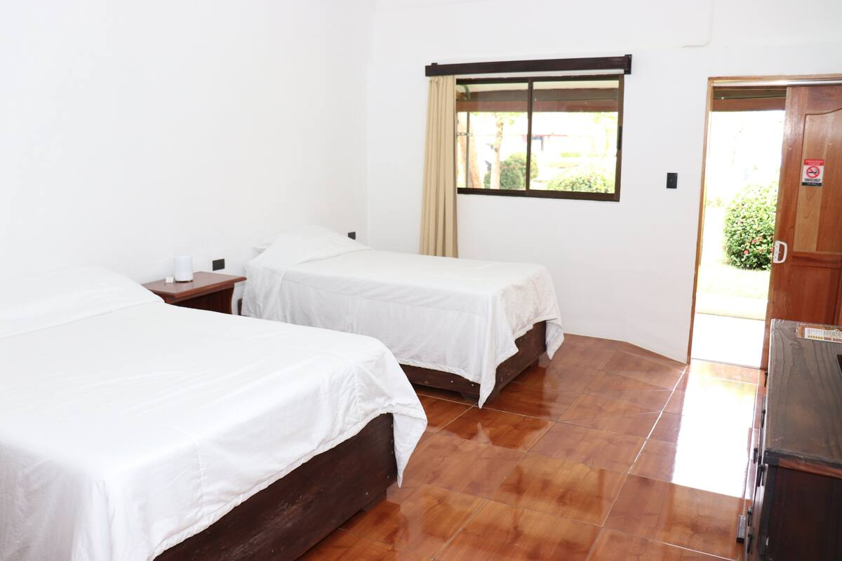 A hotel room with two single beds, a wooden wardrobe, a window with a curtain, and a tiled floor.