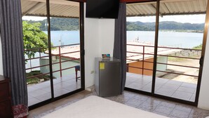 A room with a bed, a small table, and a TV. It has large windows with a view of a body of water and a distant landscape. - Hotel Samoa del Sur (Golfito)