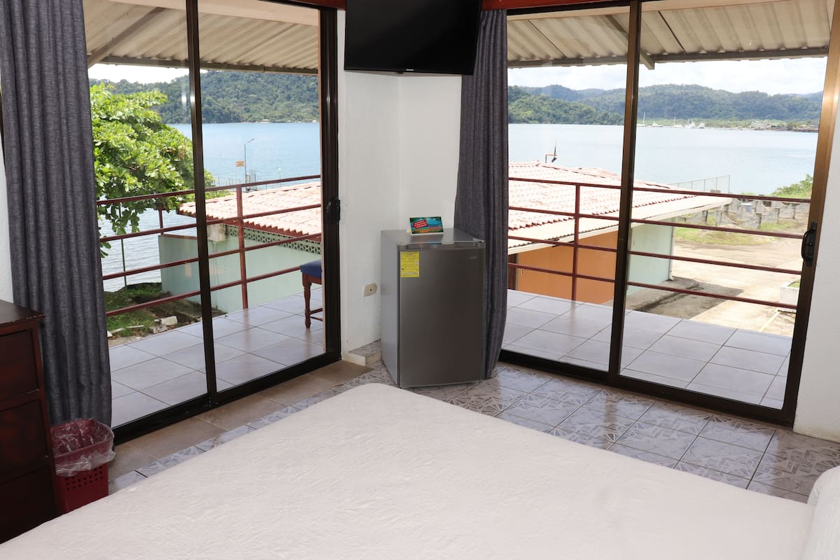 A room with a bed, a small table, and a TV. It has large windows with a view of a body of water and a distant landscape.