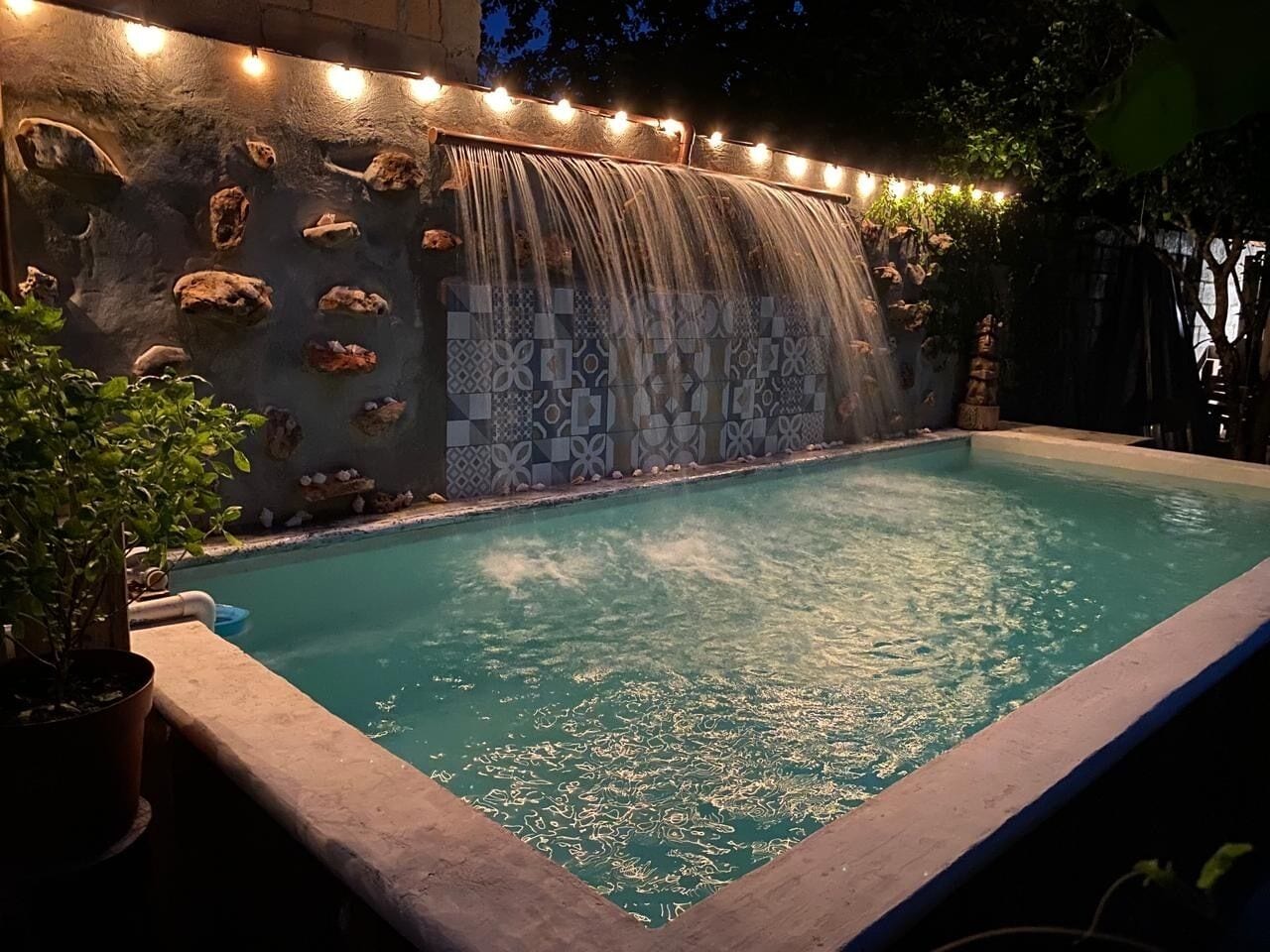 A swimming pool with a waterfall feature, surrounded by a stone wall with decorative tiles and string lights.