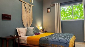 Ein Schlafzimmer mit einem Bett, einer Wanddeko aus Makramee, einem Nachttisch, einer Lampe und einem Fenster mit Vorhängen.