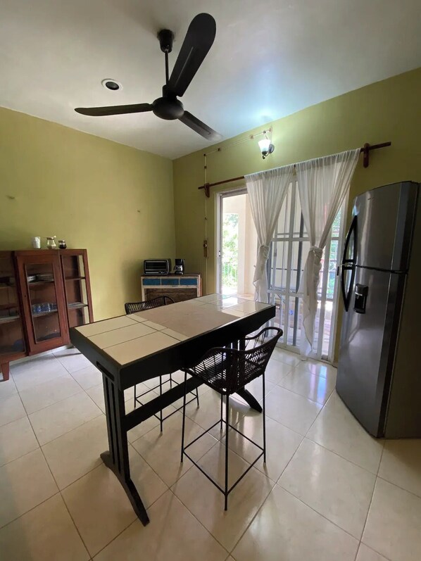 Una cocina con mesa, sillas, ventilador de techo, refrigerador y ventana con cortinas.