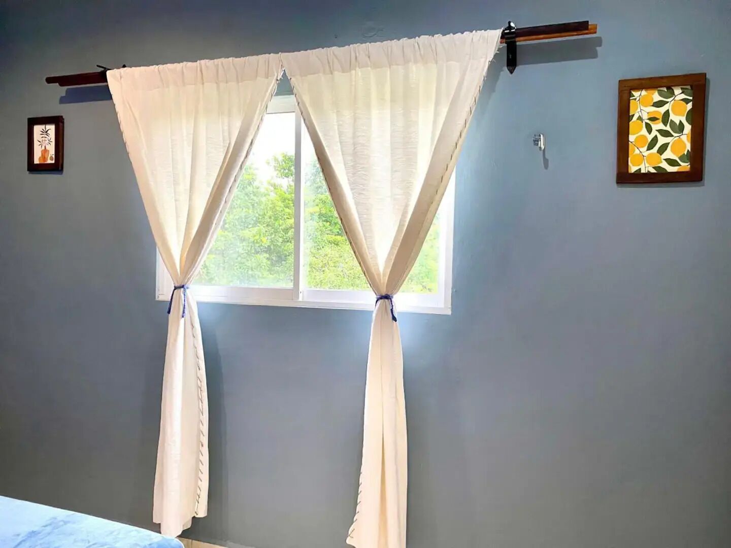 Una habitación con una ventana cubierta por cortinas blancas atadas en la parte superior. Hay dos cuadros en la pared, uno con una planta y otro con un motivo de cítricos.