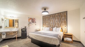 A hotel room with a large bed, a desk with a mirror, a small fridge, a hairdryer, and a clock on the nightstand.