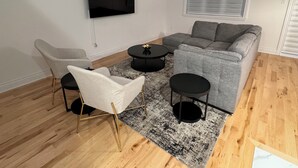 A modern living room with a grey sofa, a white armchair, and two black coffee tables. A flat-screen TV is mounted on the wall, and a window with blinds is visible.