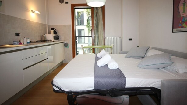 A hotel room with a bed, a small kitchenette, a dining table, and a view of the outside. - Cola Montano Apartment (Milano)