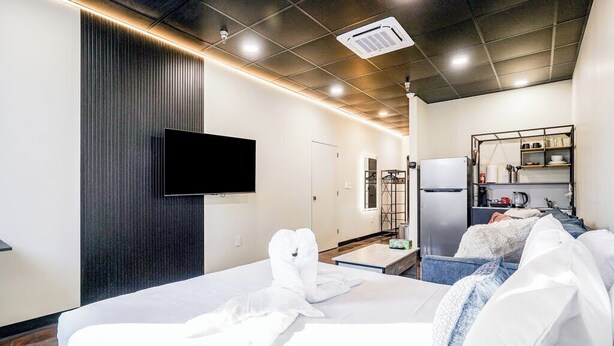 A hotel room with a bed, a flat-screen TV mounted on the wall, a kitchenette with a refrigerator, and a small seating area with a sofa and a table. - Melwood Suites Unit 106 Studio (Pittsburgh)