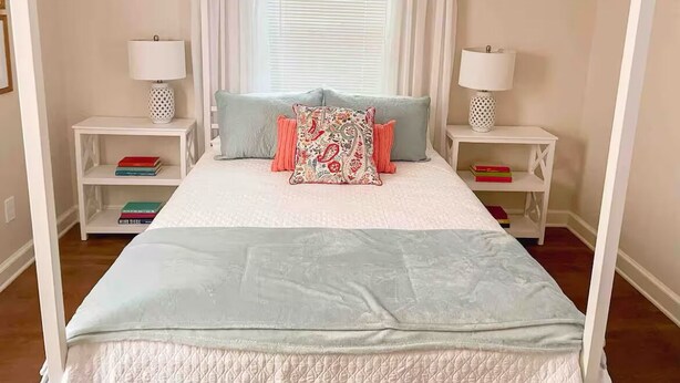 Una cama con dosel y cortinas blancas, dos mesitas de noche con lámparas y un ventanal con cortinas.