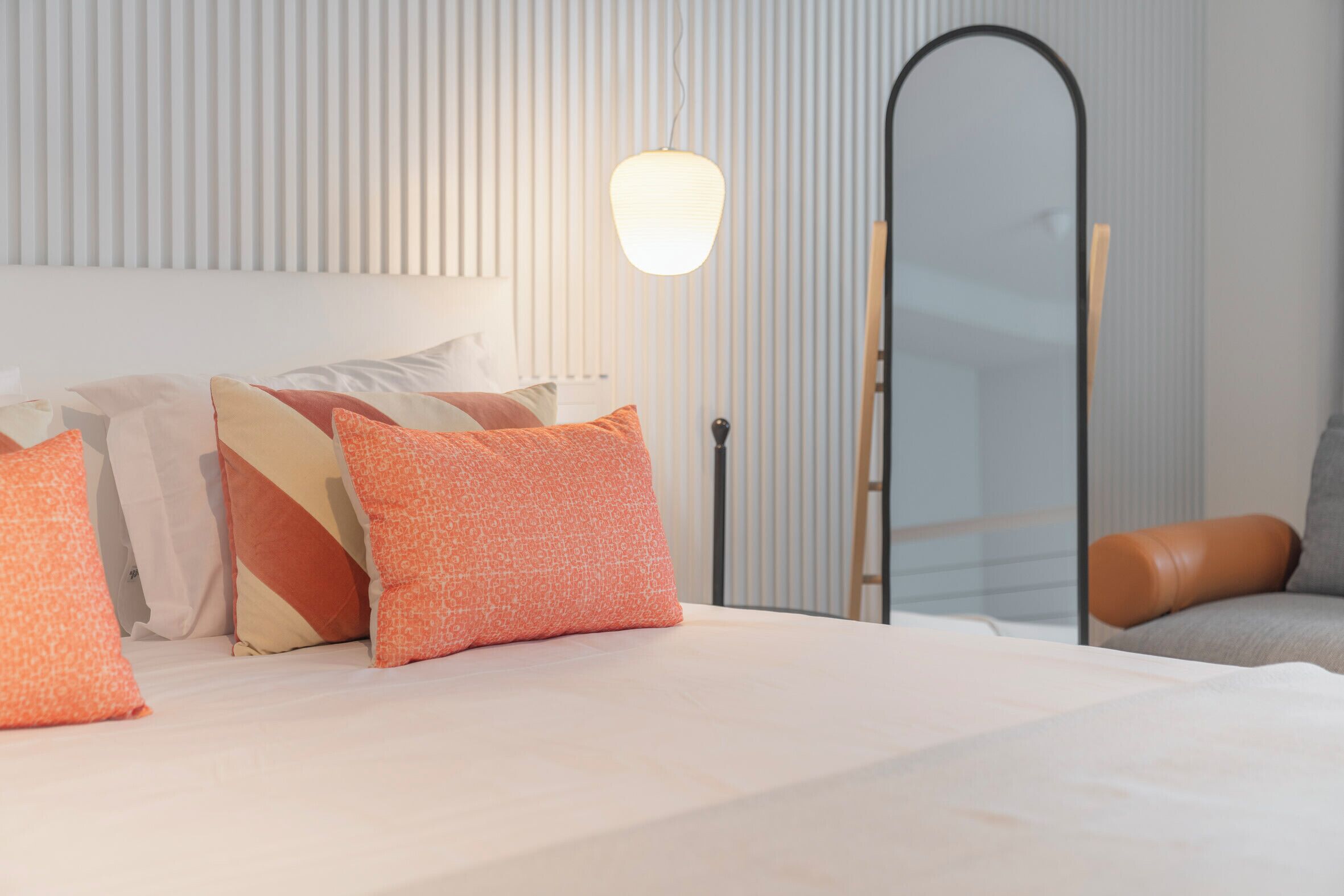 A neatly made bed with patterned pillows, a standing lamp, and a leather chair in a modern room with vertical paneling.