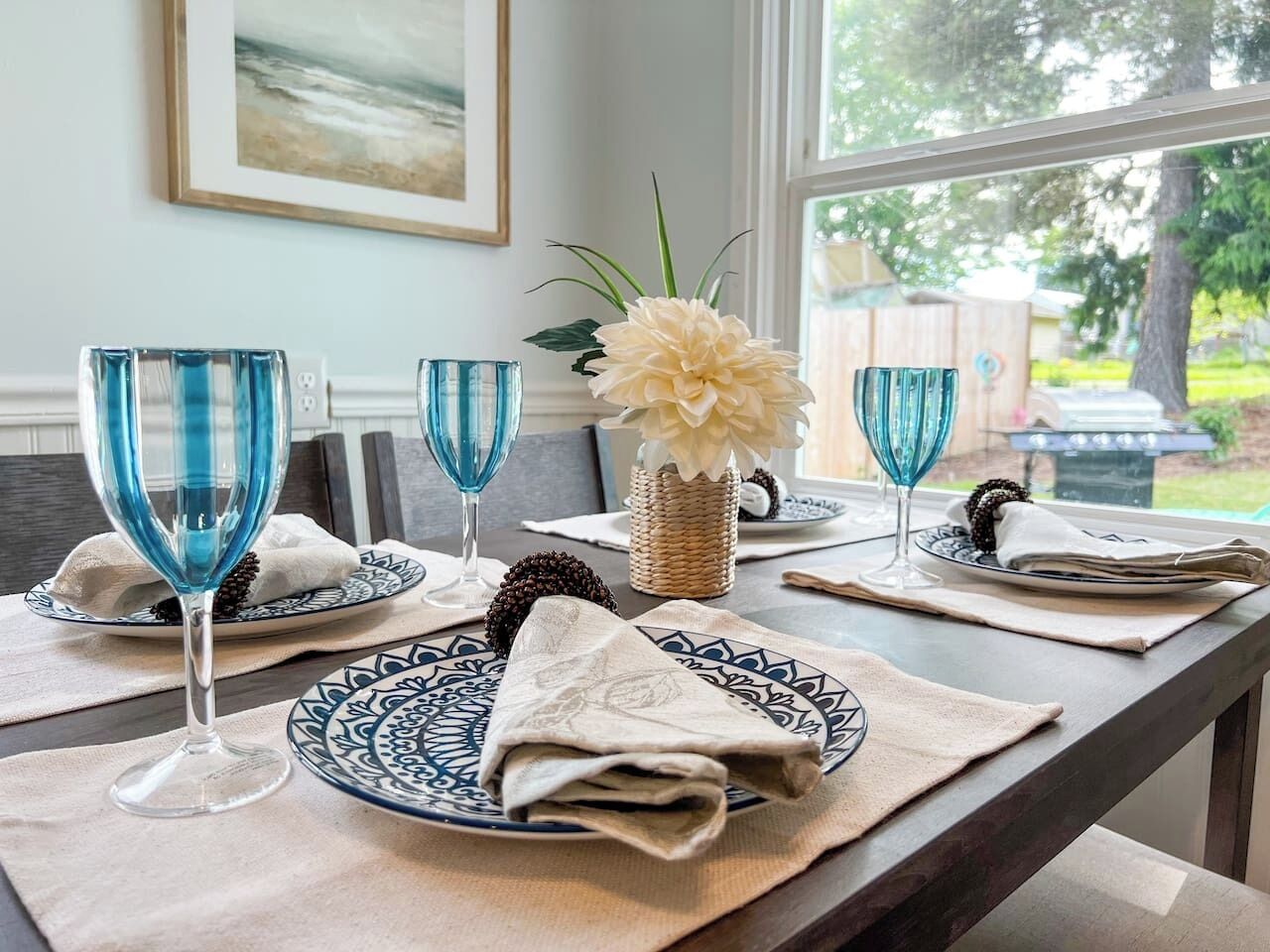 A dining table set with blue glassware, white plates, and a woven basket with a white flower centerpiece.