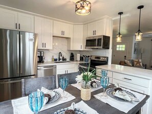 Cozinha moderna com armários brancos, eletrodomésticos de aço inoxidável e um espaço de jantar para quatro pessoas, com copos de vidro azuis e objetos decorativos.