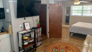 A compact living space with a flat-screen TV mounted on the wall, a bookshelf with DVDs, a small kitchen area with a sink, a bed with a patterned bedspread, and a ceiling fan. - The Unique cozy studio (Winter Haven)