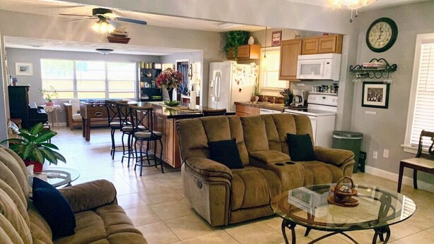 Ein großzügiges Wohnzimmer mit einer Couch, einem Relaxsessel und einem Glastisch. Im Hintergrund befindet sich eine Küchenzeile mit einer Mikrowelle und einem Kühlschrank.
