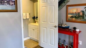 A hallway with a white door leading to a bathroom, a red console table with nautical decorations, and a framed painting on the wall.