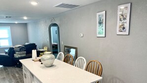 Una cocina con una isla grande, sillas de madera y una mesa de comedor. Hay cuadros en la pared y una ventana con persianas.
