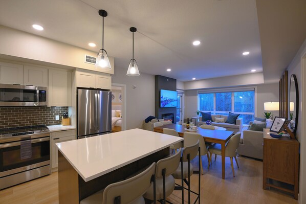 A modern kitchen with stainless steel appliances, a dining area with a large island, and a living room with a sofa and TV.